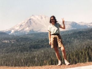 A young women with the vicotroy sign wearing sunglasses, T-shirt and shorts standing on the top of a mountain