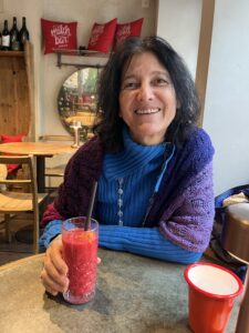 A dark haired women, smiling with a blue pullover holding a berry smoothie in her hand
