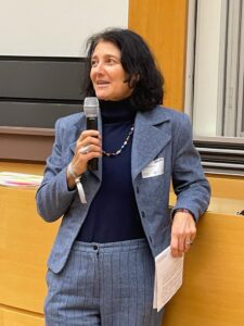 A darkheard women in a blue suit standing with a microphone in her right hand and speaking to the audiance