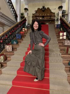 a dark haired women, smiling, standing on a red carpet in a beautiful staircase with lots of Christmas presents on the stairs