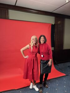 Two women standing infront of a red background. One woman is blond and wearing a red dress. The other woman is wearing a red pullover and a black skirt.
